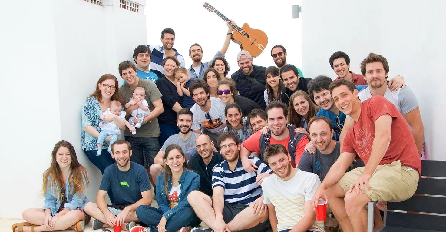 Large group photo of the Tarmac team gathered on a sunny terrace, some holding babies and a guitar